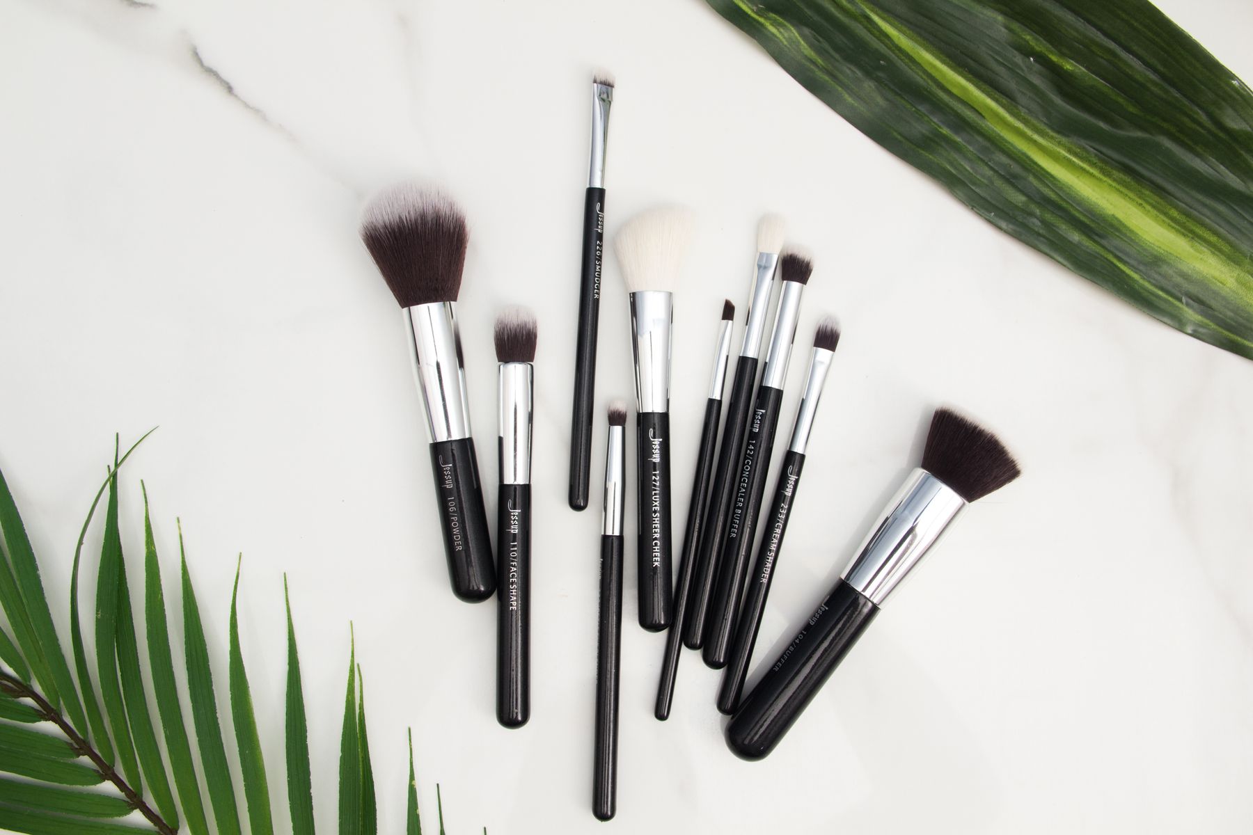 Assorted makeup brushes arranged on a white surface beside green palm leaves