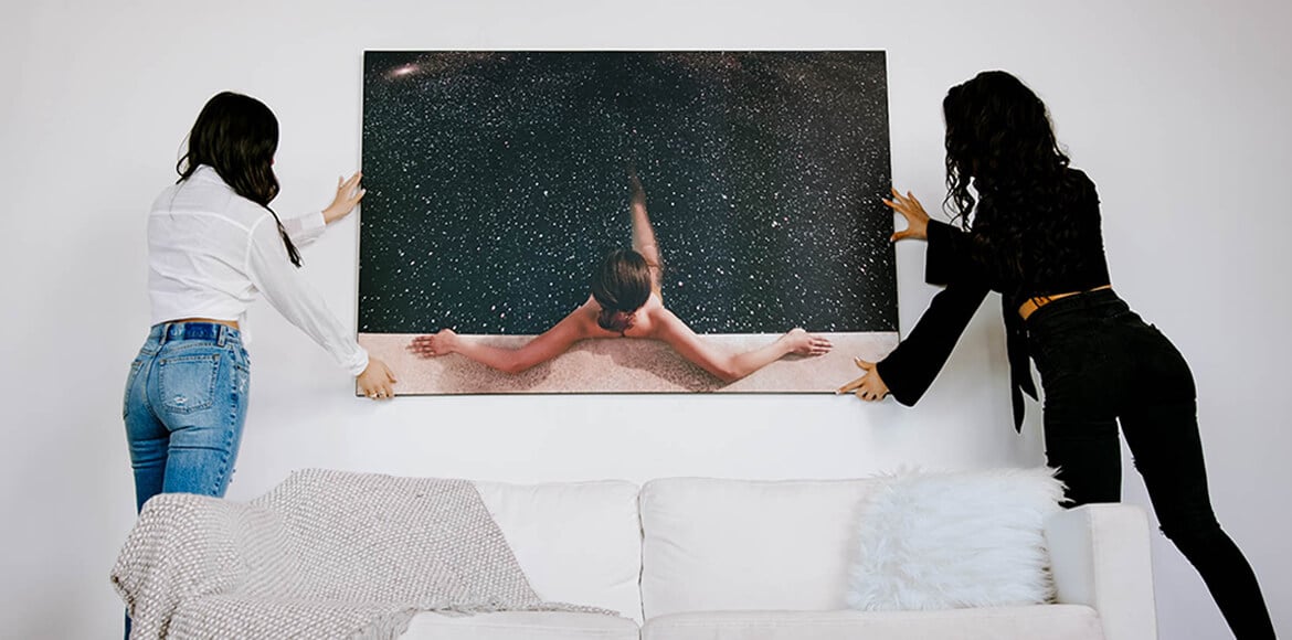 Two people hanging an oversized surreal canvas above a white sofa