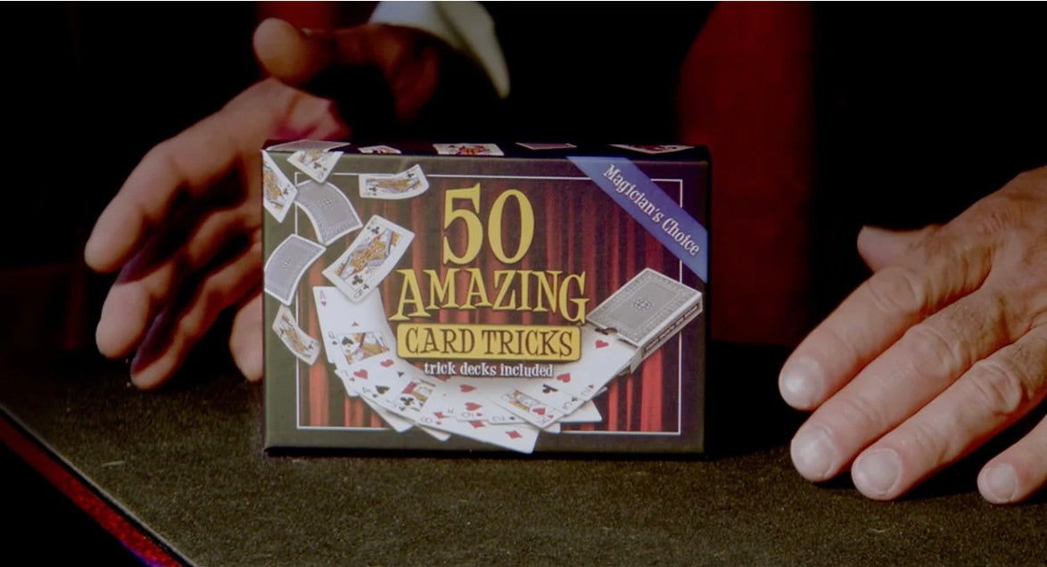 Hands presenting a boxed 50 Amazing Card Tricks kit on a dark tabletop