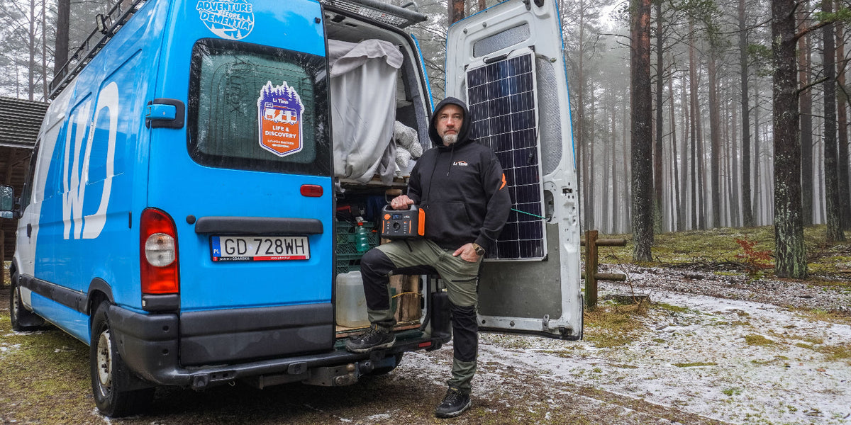 Man holding a portable power station beside a blue camper van in a wintry forest.