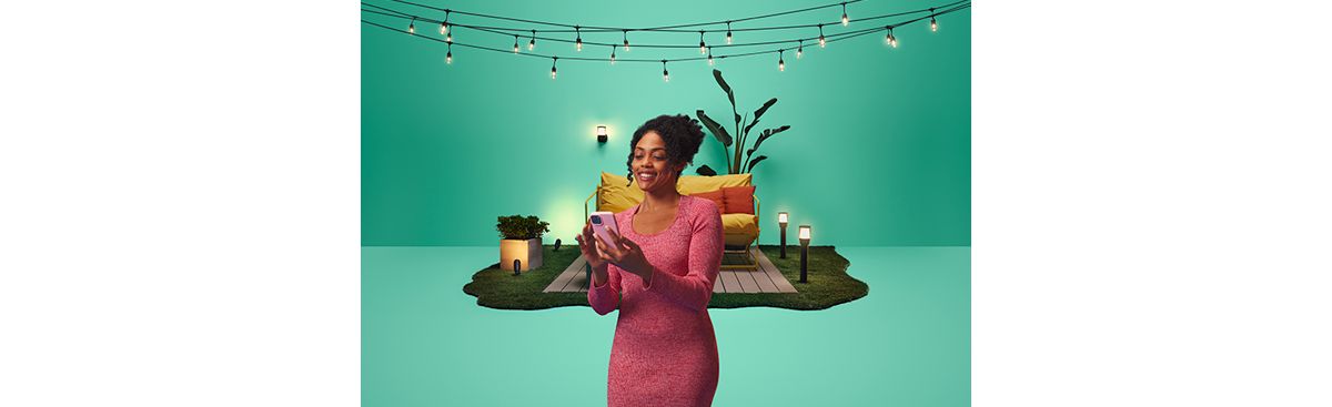 Woman using a phone in front of a staged patio lit by string lights and outdoor lamps