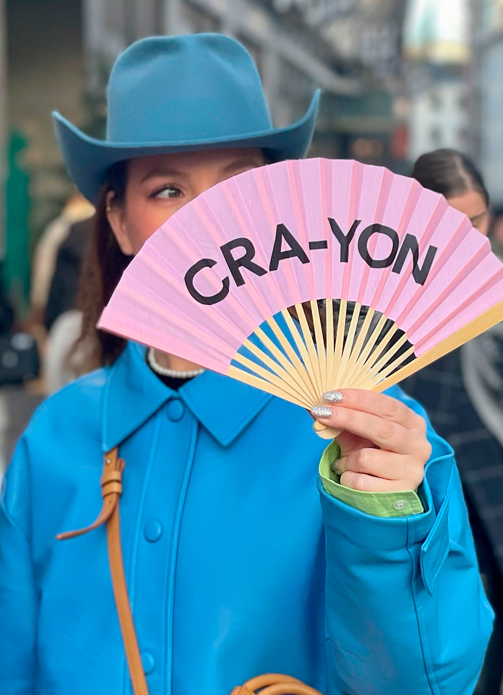 Fashion model in a turquoise coat and cowboy hat holding a pink CRA-YON fan on a city street
